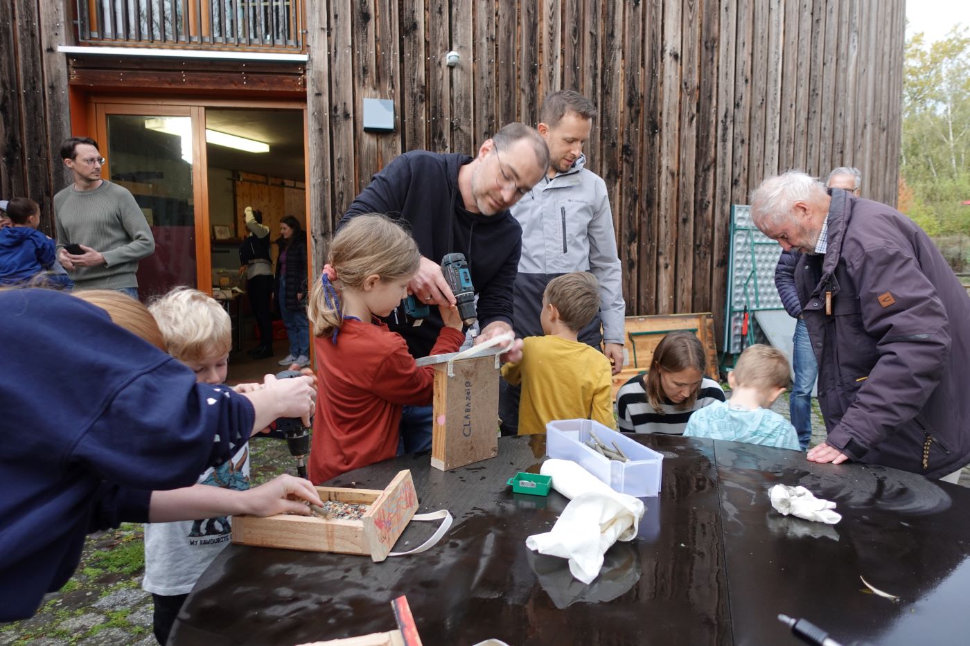 Kinder und Erwachsene arbeiten im Freien gemeinsam mit Holzrahmen, Vogelfutter und Werkzeug.